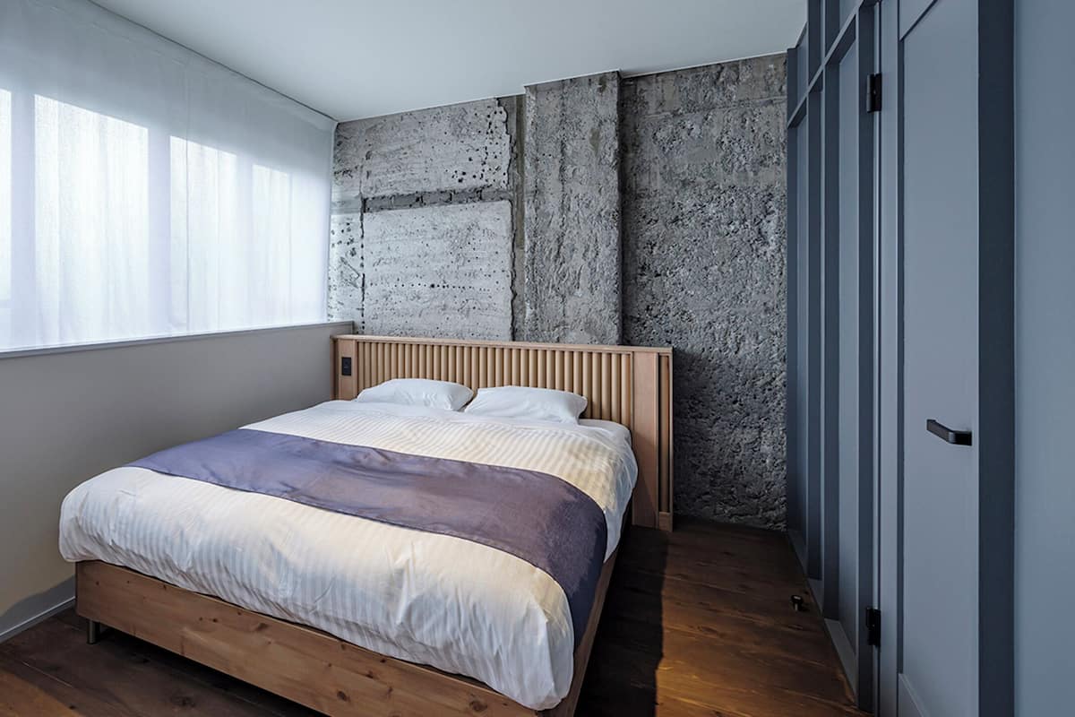 A modern bedroom with a wooden bed, a concrete wall, and a large window with sheer curtains.