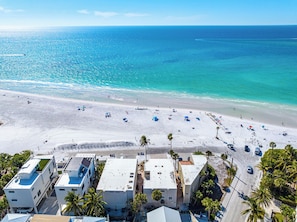 An aerial view of a beach with white sand, clear blue water, and a few buildings in the foreground.