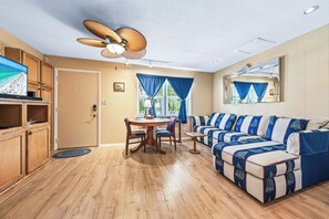 A room with a wooden floor, a dining table with chairs, a sofa, a television, and a ceiling fan.