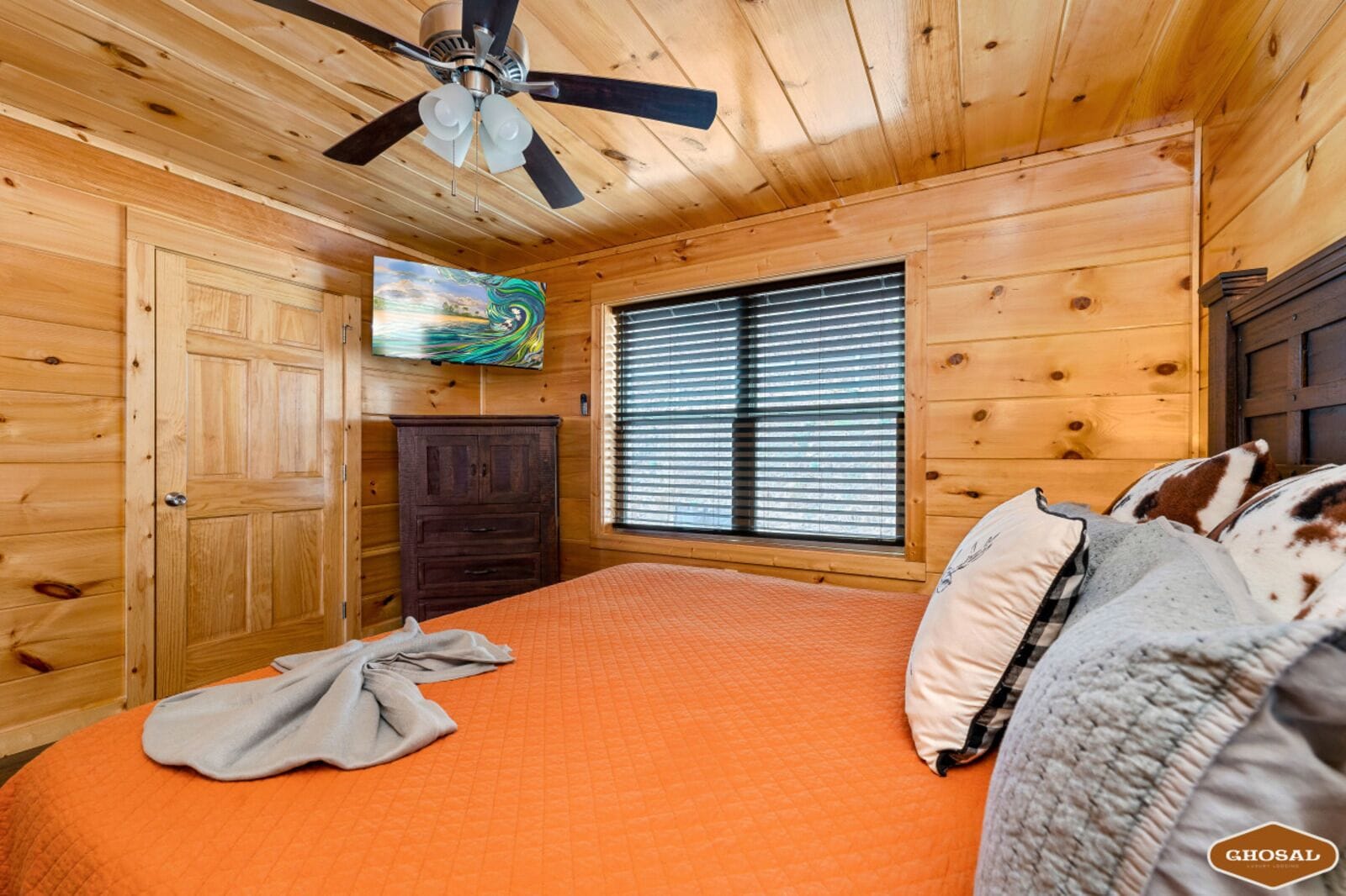 A wooden cabin bedroom with a bed, a ceiling fan, a window with blinds, and a TV on the wall.