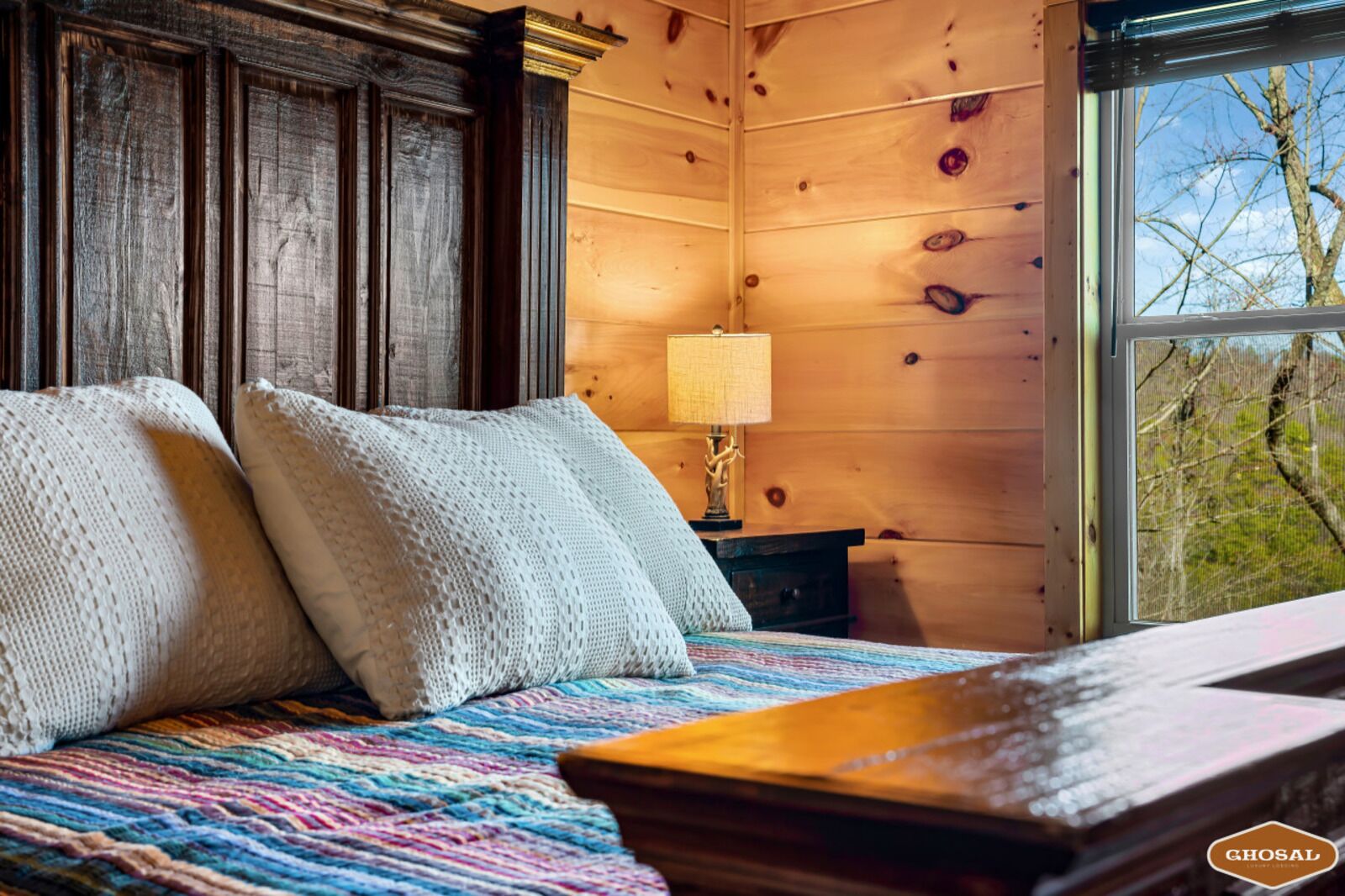 A cozy room with wooden walls, a bed with two white pillows, a colorful blanket, and a window with a view of trees.