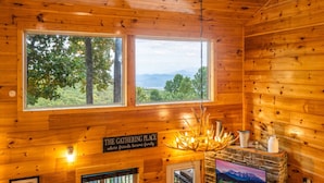 Een houten hut interieur met een groot raam dat uitkijkt op een bos en bergen. Een bordje staat erop: "The Gathering Place where friends become family.".