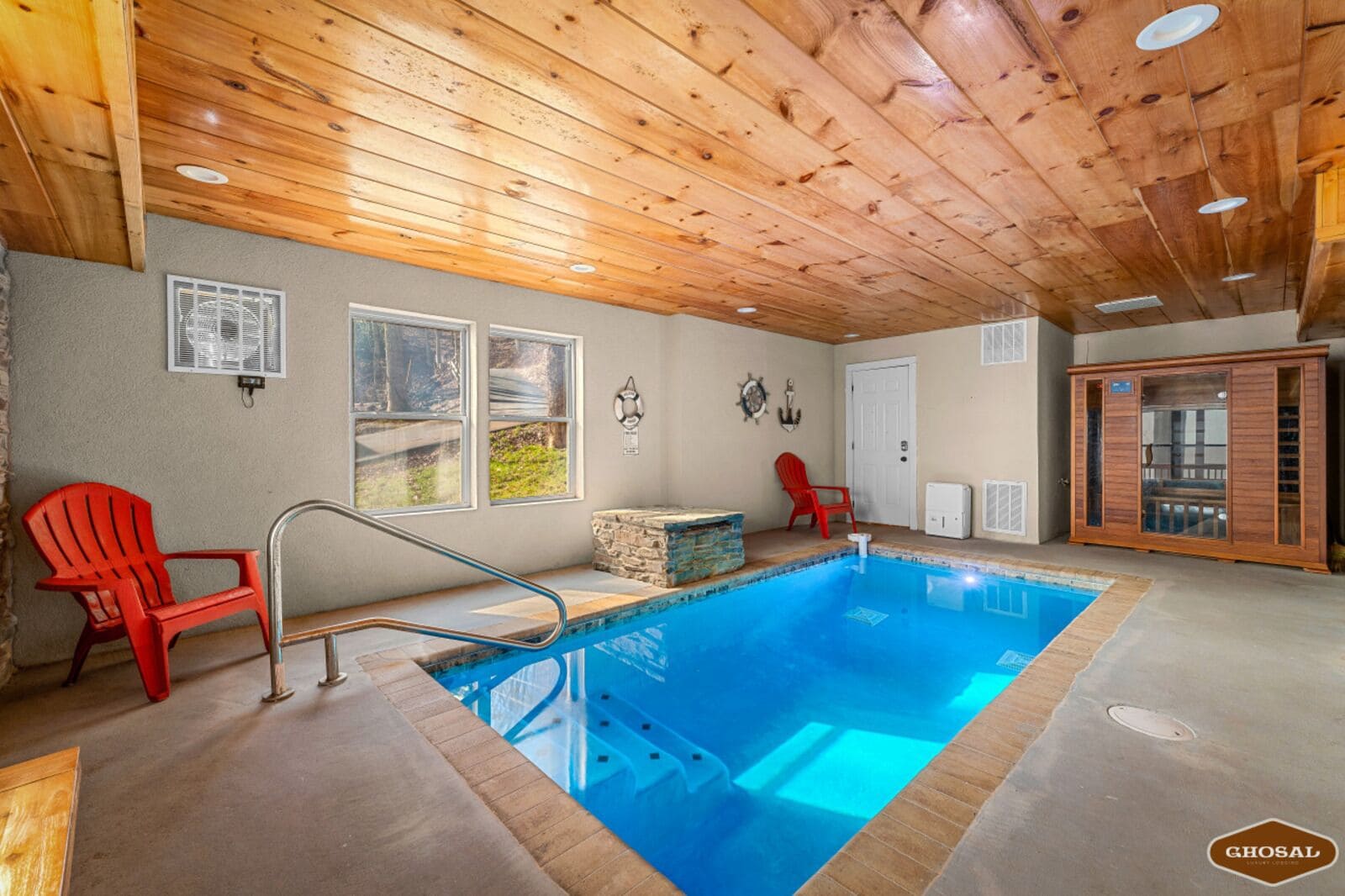 A pool area with a wooden ceiling, two red chairs, a stone feature, and a glass cabinet.