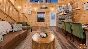 Een gezellig blokhutinterieur met houten inrichting, een stenen open haard, een eethoek en een bord met de tekst 'The Gathering Place'.