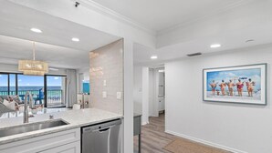 A modern kitchen with a dishwasher, a large window overlooking the ocean, and a painting on the wall. - Emerald Towers 305 (Destin)