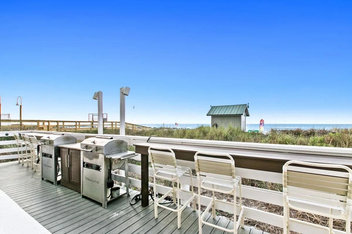 Een houten terras met barbecuegrills en witte metalen stoelen, met uitzicht op een strand.