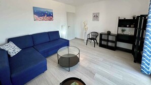 En moderne stue med en blå sofa, et glasbord, en sort lænestol, et sort reolsystem og et maleri af et bybillede.