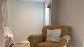 A beige armchair with a matching ottoman and a decorative pillow in a room with tiled flooring and a window with blinds. - Beachfront Bliss: Water Front, Steps to Sand, Minutes from Dining & Attractions! (New London)