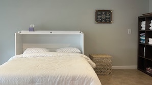 A bedroom with a white bed, a small bedside table, a woven stool, a dark wooden wardrobe, and a patterned rug on the floor. - Beachfront Bliss: Water Front, Steps to Sand, Minutes from Dining & Attractions! (New London)
