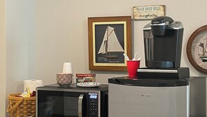 A kitchen with a microwave, a coffee maker, a framed picture, and a basket with items. - Beachfront Bliss: Water Front, Steps to Sand, Minutes from Dining & Attractions! (New London)