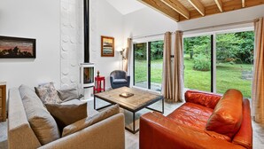 A modern living room with a wooden ceiling, a large window with a view of greenery, a sofa, a coffee table, and a chair. - Nordic Eco House - Black Mountain (Black Mountain)
