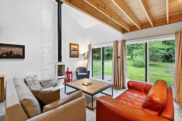 A modern living room with a wooden ceiling, a large window with a view of greenery, a sofa, a coffee table, and a chair.