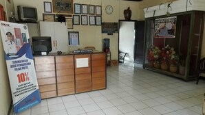Zona de recepción de un hotel con un mostrador de madera, certificados enmarcados en la pared, un reloj y un arreglo floral.