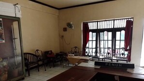 Una habitación con un banco de madera, una mesa con un letrero que dice 'PENUH', un ventilador, una ventana con barrotes y cortinas rojas, y un soporte para bicicletas afuera.