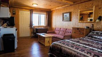 A cozy cabin room with a wooden ceiling, a bed with a forest-themed blanket, a red and white checkered sofa, a wooden dresser, and a desk with a computer and a lamp.