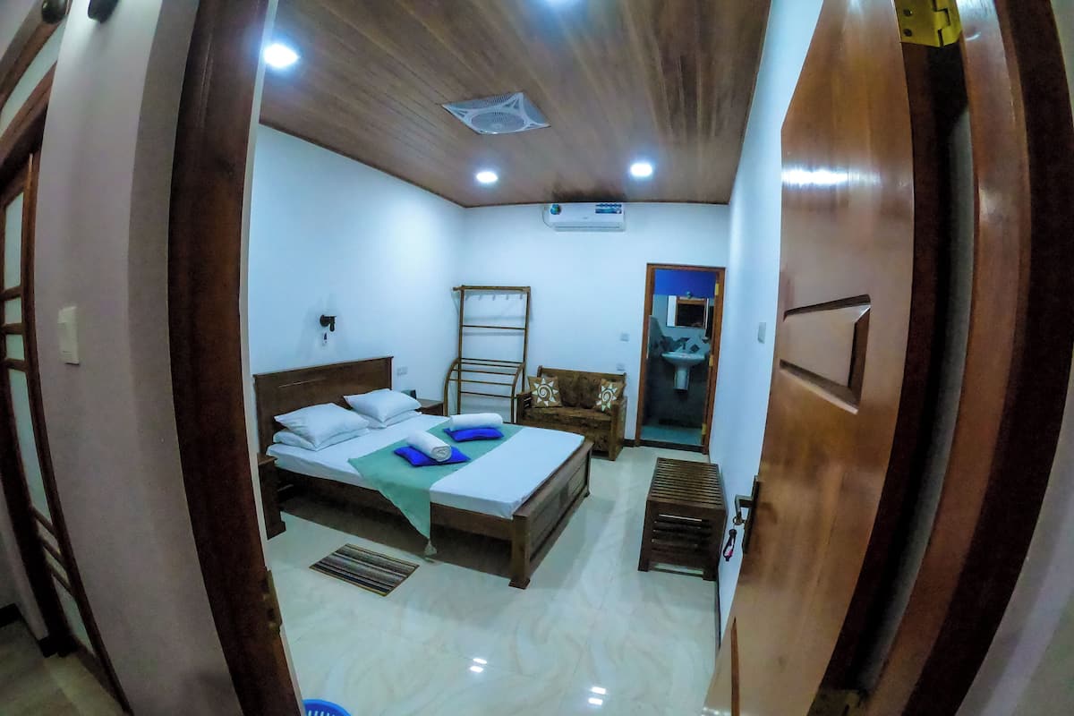 A bedroom with a bed, a wooden headboard, a small table, a blue basket, and a bathroom visible through an open door.
