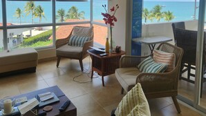 Ein Zimmer mit Meerblick, ausgestattet mit Deckenventilator, großen Fenstern mit Jalousien, Korbmöbeln und einem kleinen Tisch mit einer Vase und Fernbedienungen.