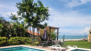 Ein Swimmingpool mit einer Pergola und Liegestühlen, umgeben von tropischer Vegetation und Blick auf den Ozean.