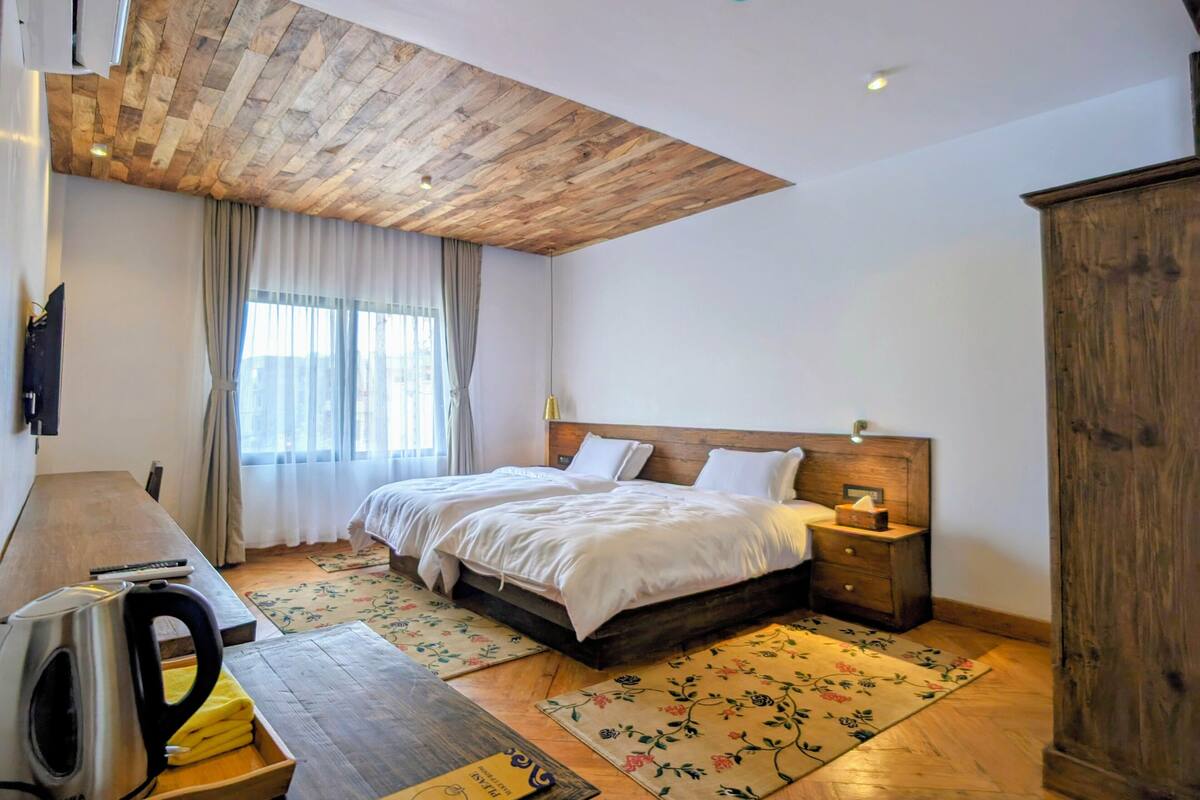 A bedroom with a wooden ceiling, a bed with white bedding, a nightstand, a wooden cabinet, and a kettle on a table.