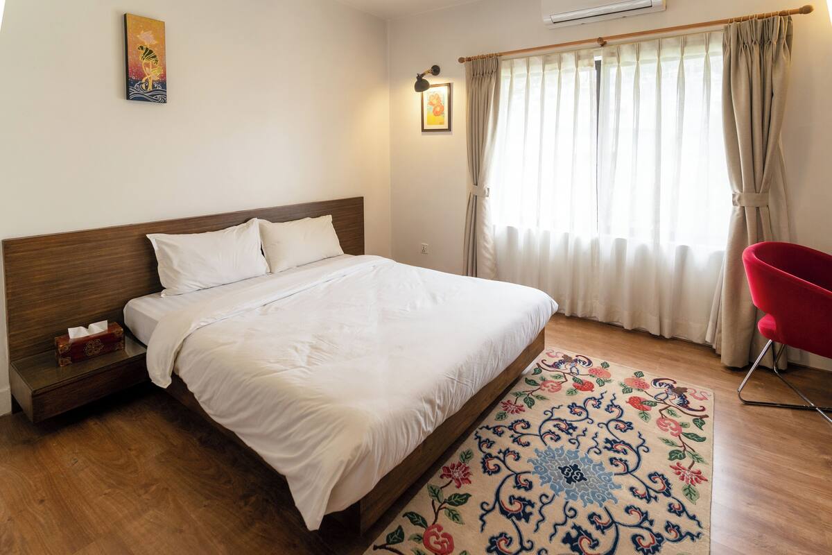A neatly made bed with white linens, a wooden headboard, a bedside table with a tissue box, a red chair, and a patterned rug on a wooden floor.