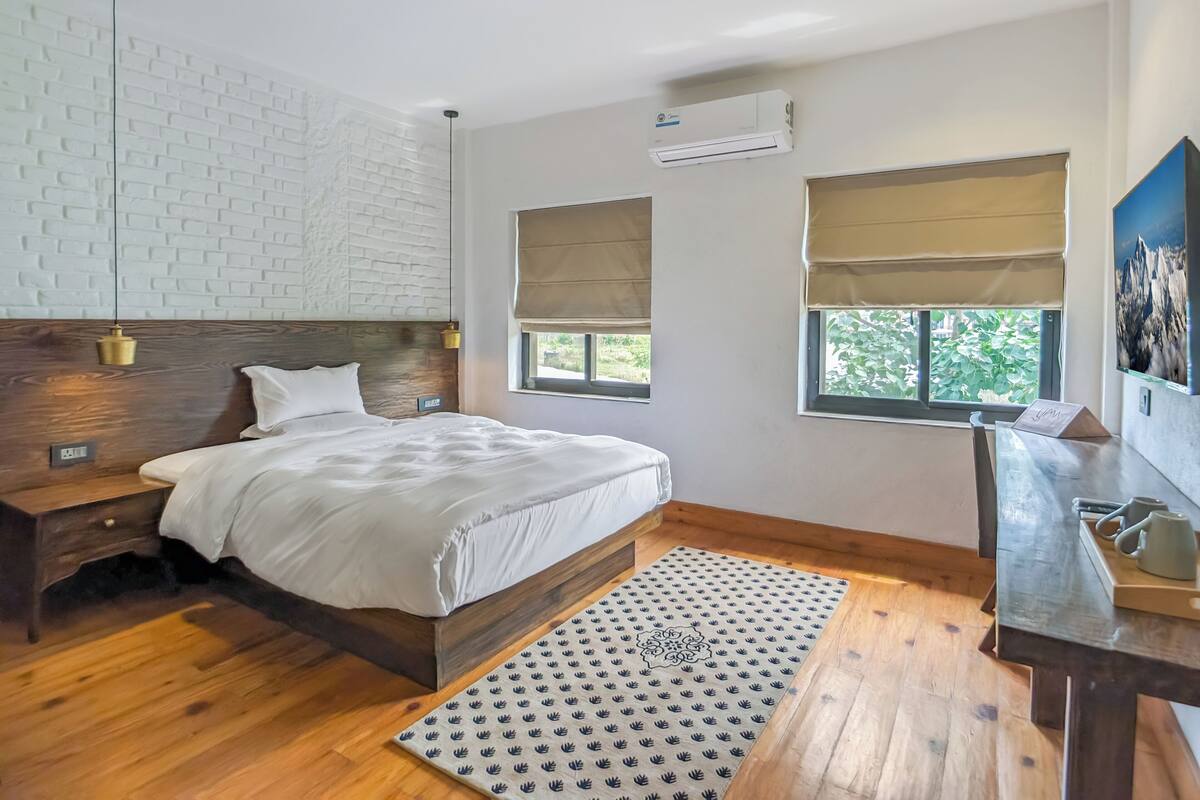 A modern bedroom with a wooden bed, white bedding, a patterned rug, and a view of greenery through the windows.