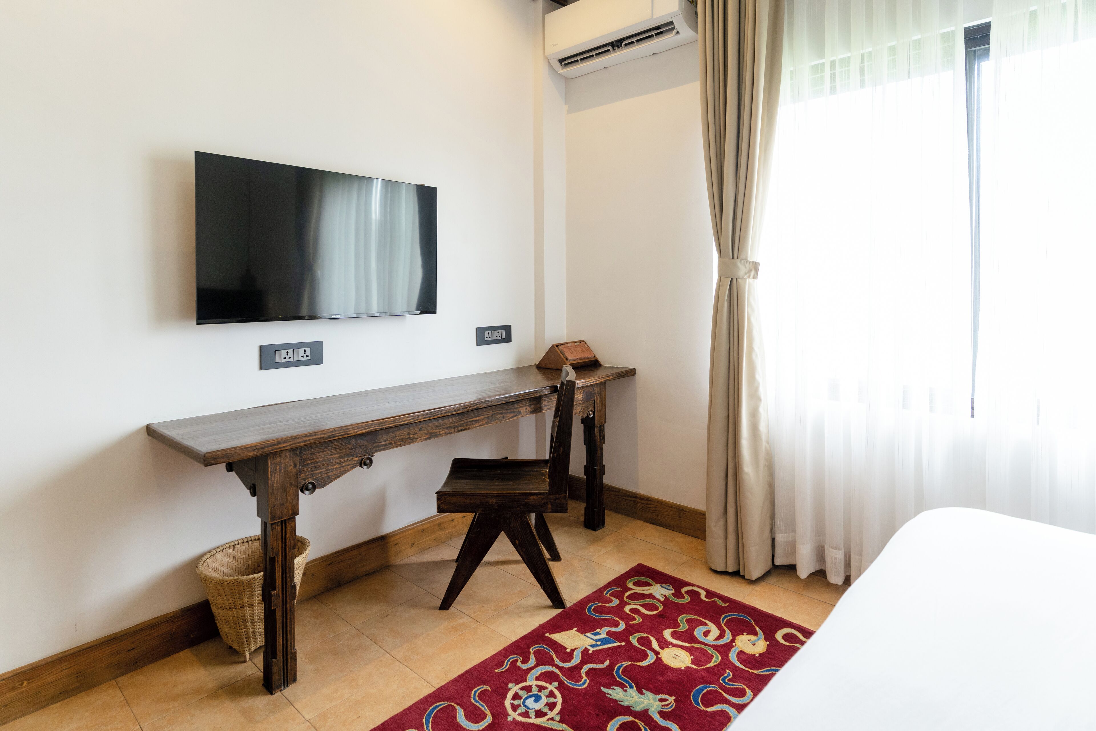 Camera d'albergo con televisore a schermo piatto montato a parete, una scrivania in legno con una sedia, un tavolino con una lampada, un tappeto rosso decorato e un'ampia finestra con tende leggere.