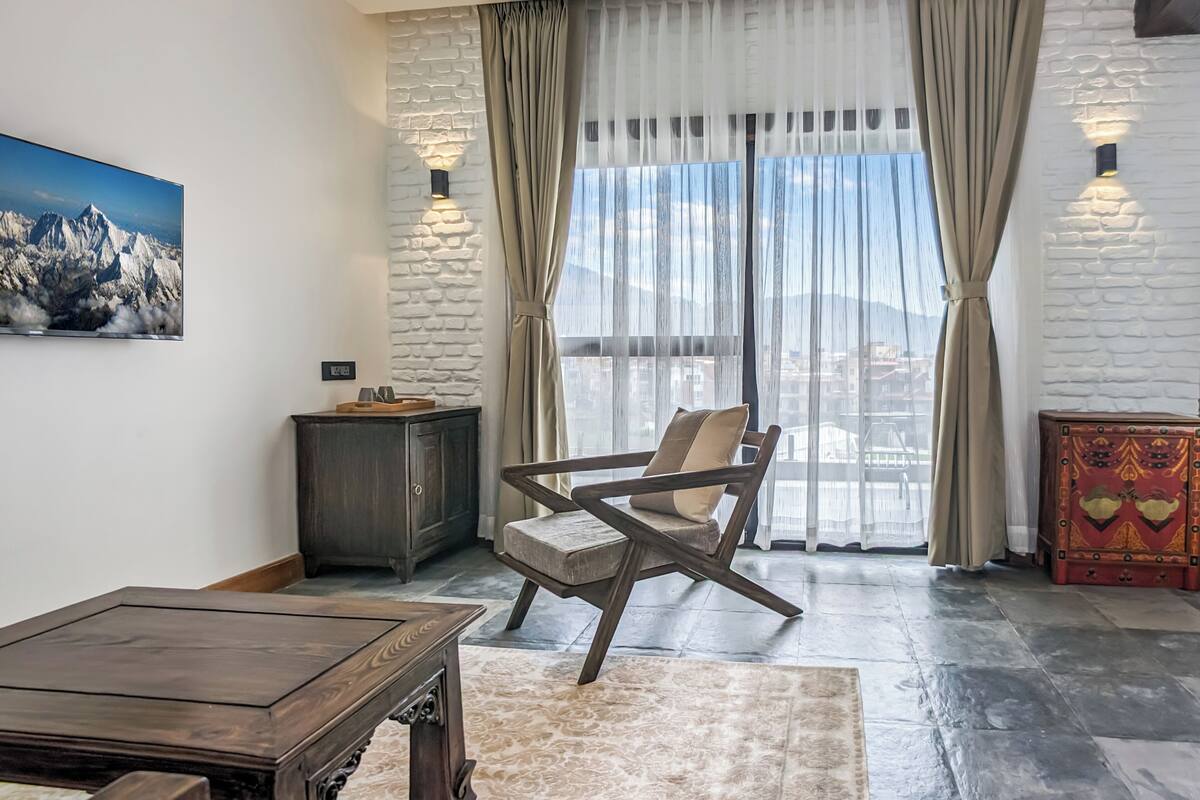 A modern living room with a wooden coffee table, a cushioned armchair, a small cabinet, and a large window with sheer curtains.