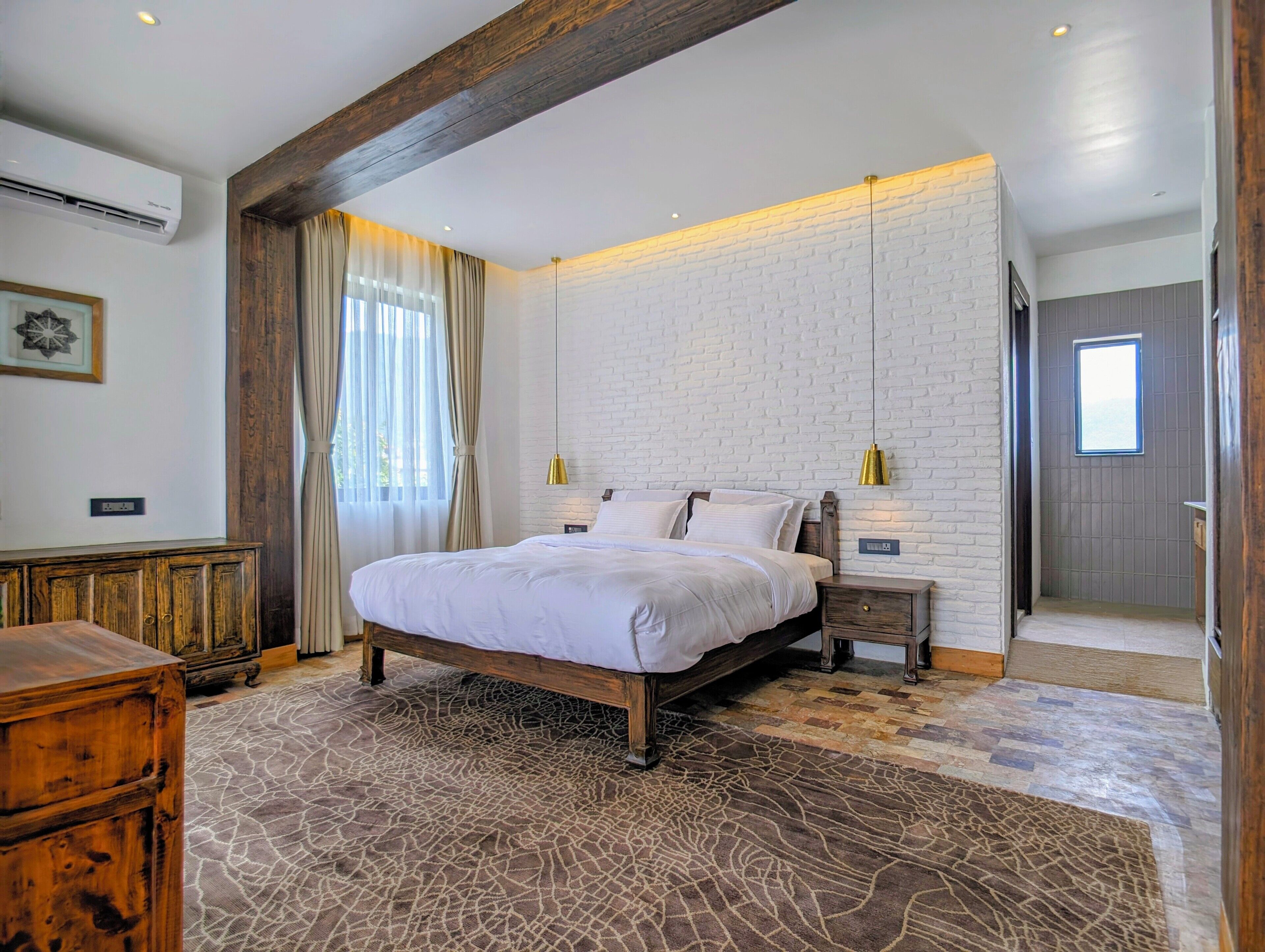 A bedroom with a large bed, wooden headboard, two bedside tables, a patterned rug, a white brick wall, and a window with curtains.