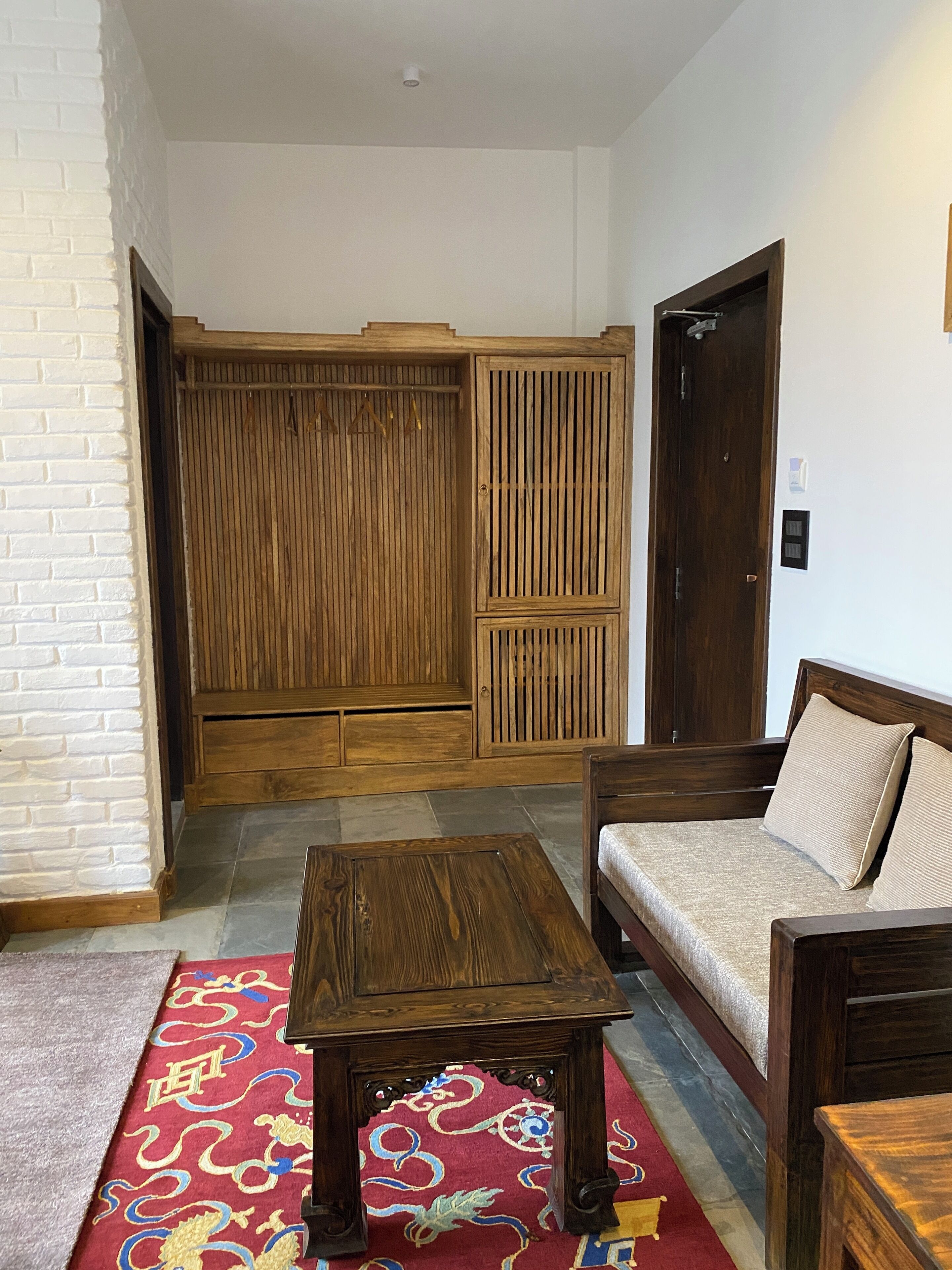 A room with a wooden wardrobe, a sofa with cushions, a coffee table, and a patterned rug.