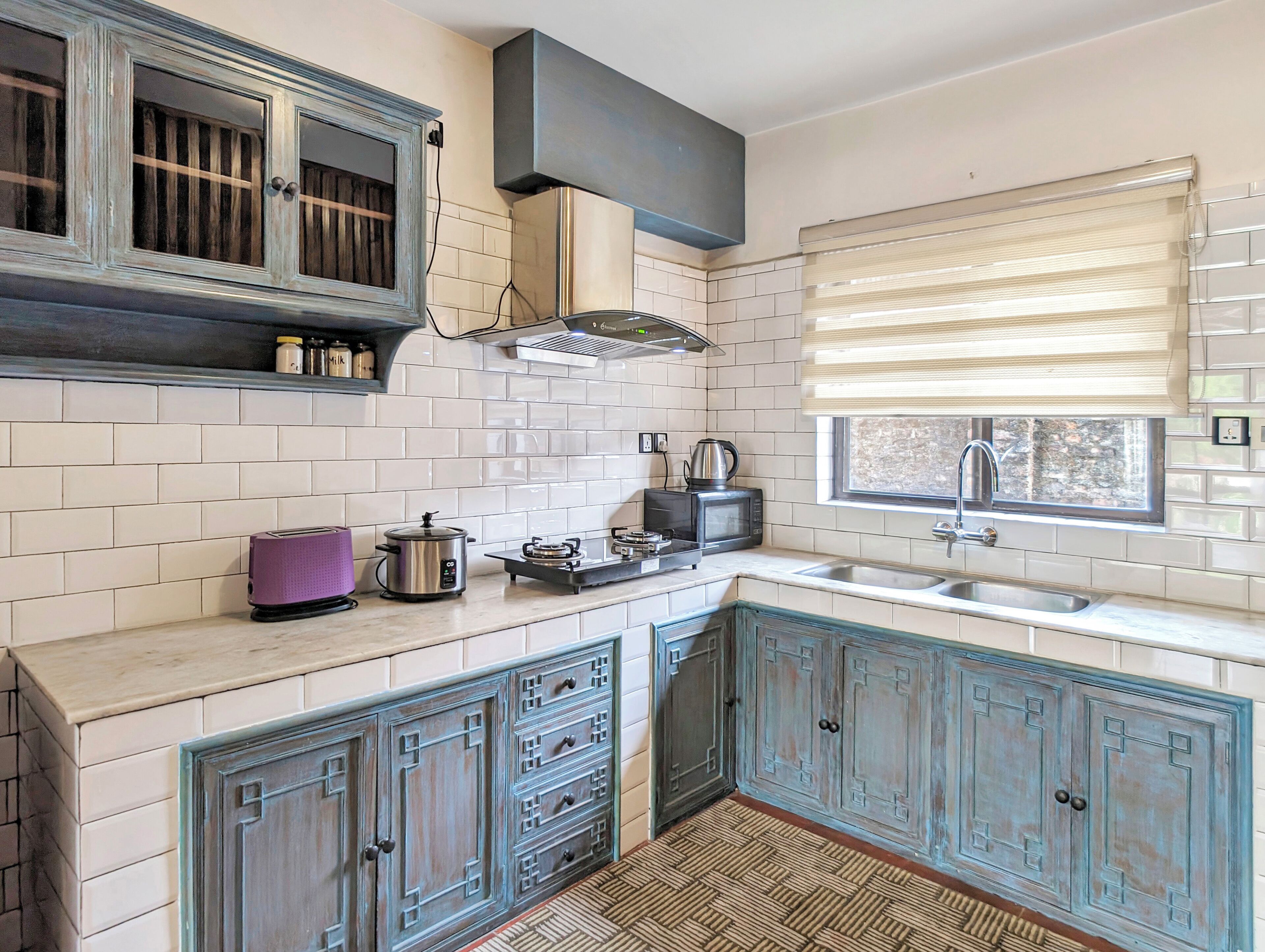 Une cuisine avec des murs recouverts de carreaux blancs, une fenêtre avec des stores, un évier, une cuisinière et des armoires à l’aspect rustique.