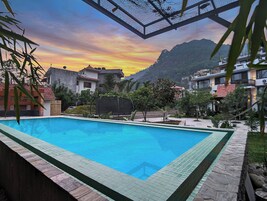 Piscina con borde de azulejos, rodeada de edificios y follaje, con montañas al fondo.