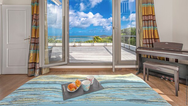 Een kamer met een grote schuifdeur die leidt naar een balkon met uitzicht op zee. Er staat een bed met een gestreept dekbed, een klein tafeltje met een schaal croissants, en een piano.