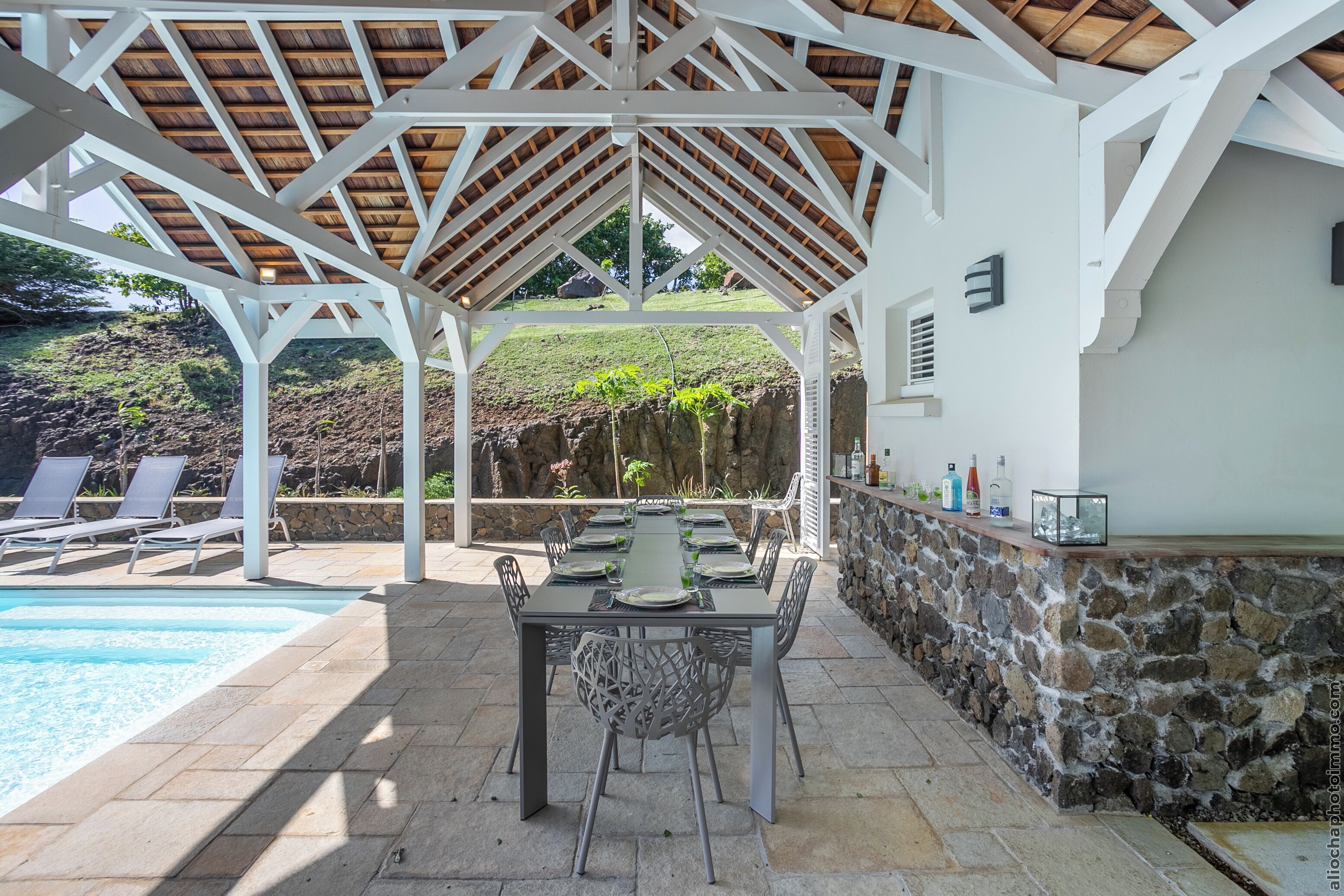 A covered outdoor dining area with a pool, stone wall, and lounge chairs.