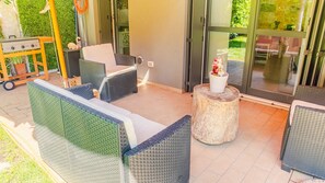 Un espace patio aménagé avec des meubles en osier, une petite table ornée d’un vase de fleurs et un chariot en bois muni d’un barbecue.