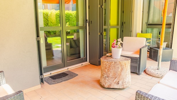 A patio area with a sliding glass door leading to a garden, a round wooden table with a potted plant, and wicker furniture. - Lugana Lovers - Italian Homing (Sirmione)