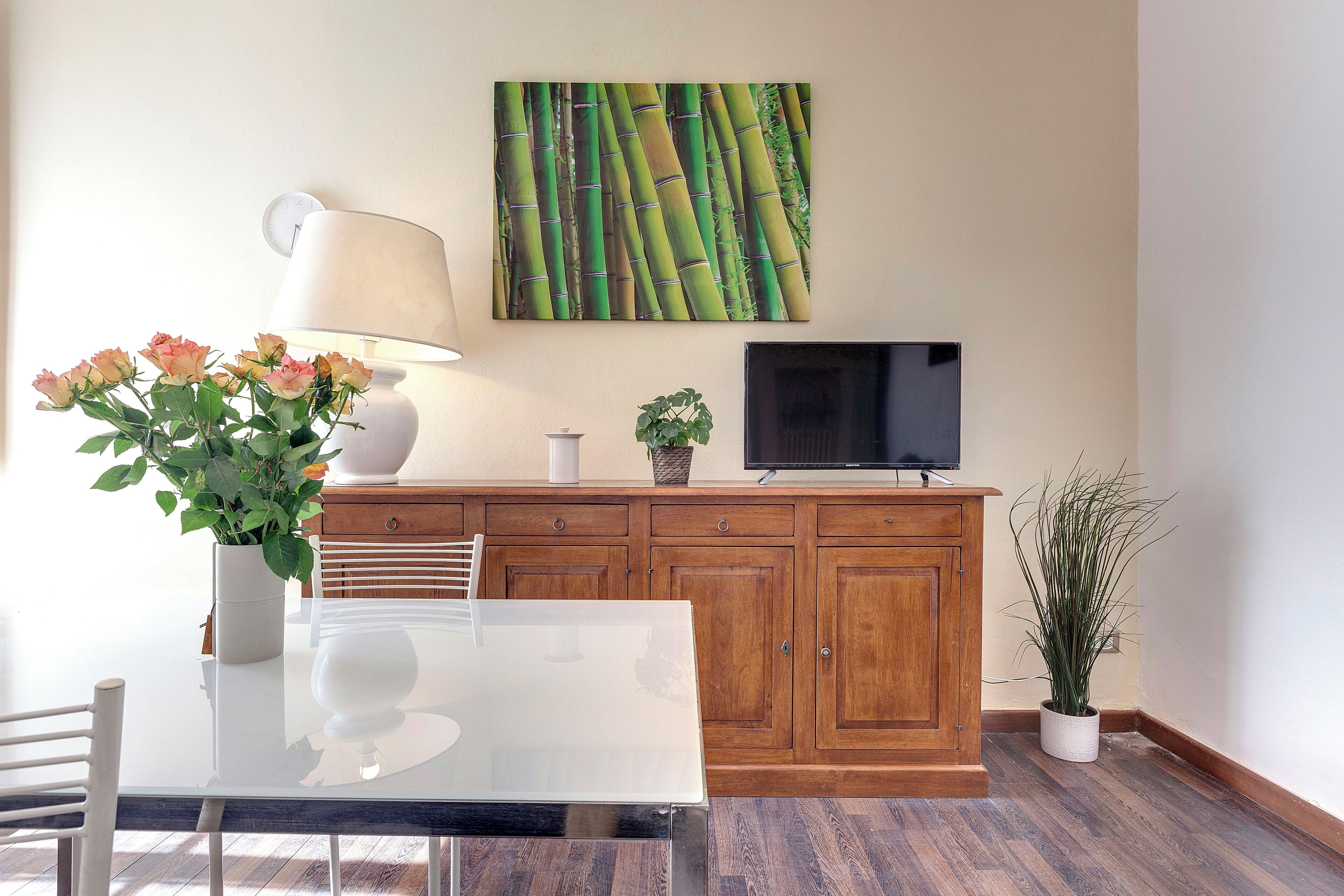 A dining area with a glass table, chairs, a lamp, a potted plant, a television on a wooden cabinet, and a wall-mounted clock.