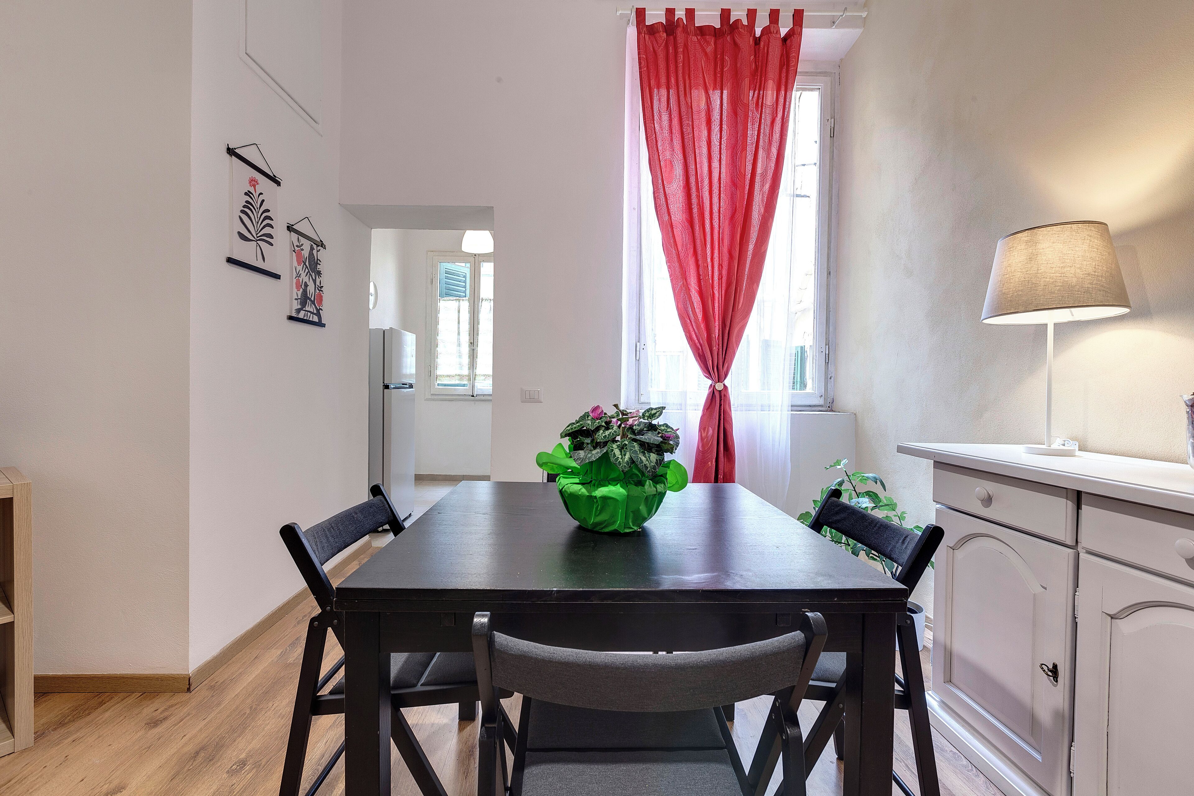A dining area with a table, chairs, a green plant, a red curtain, and a lamp.