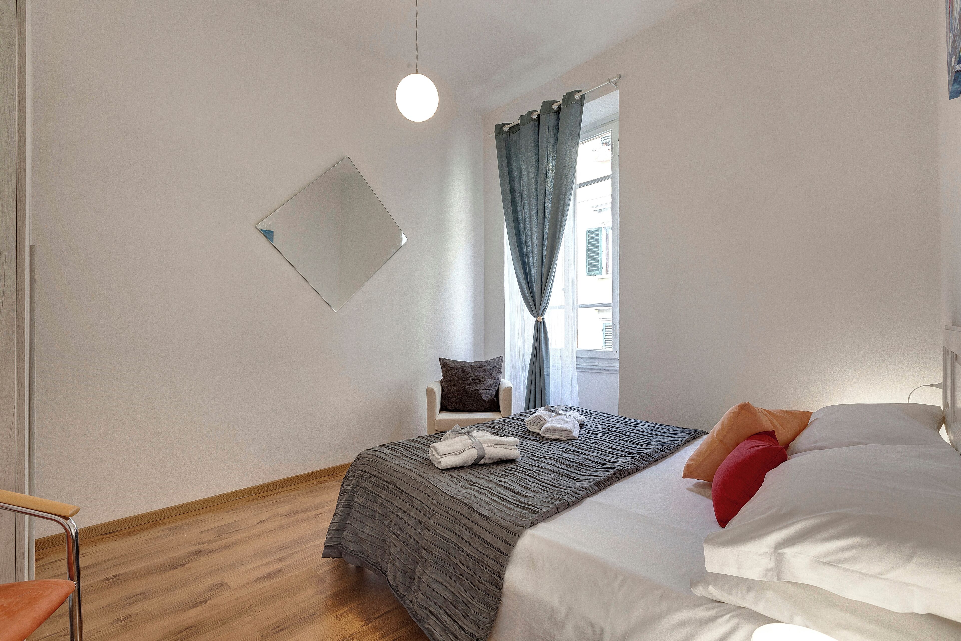 A bedroom with a bed, a chair, a window with curtains, and a hanging light fixture.