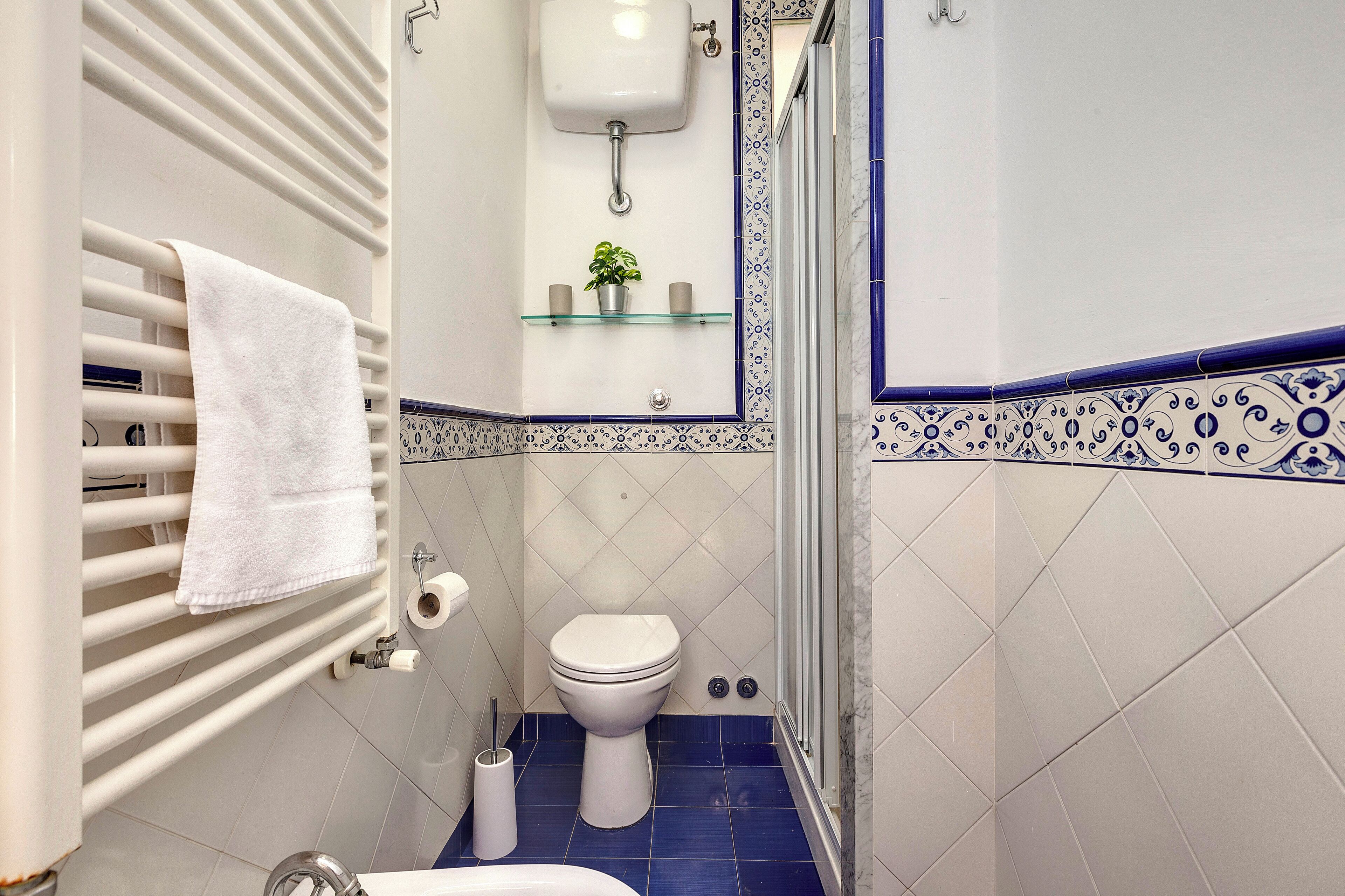 A bathroom with a white radiator, a white toilet, a glass shelf with a plant, and a shower area.