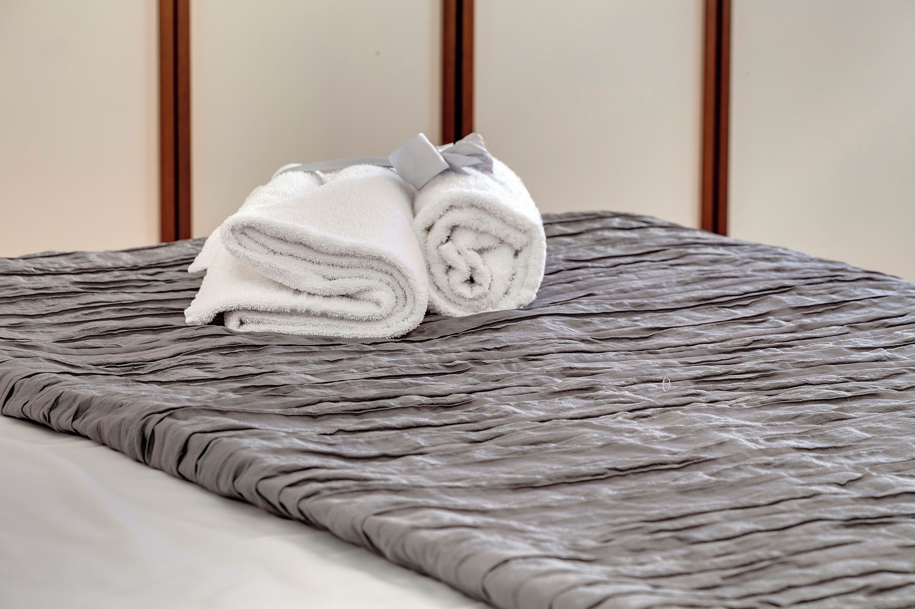 Een net opgemaakt bed met een grijs geweven sprei en een opgerolde witte handdoek met lint.