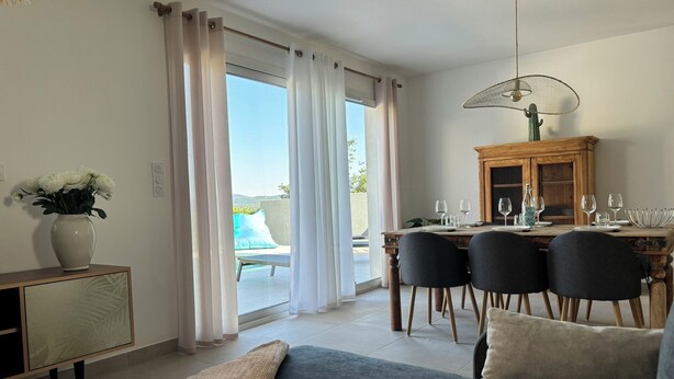 A modern living room with a sofa, a dining table set for four, and a view of the outdoors through large windows. - Maison au Calme Avec Piscine (SAINT MICHEL D'EUZET)
