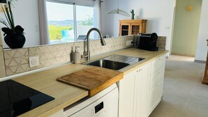 A modern kitchen with a large island, a built-in dishwasher, and a view through the window. - Maison au Calme Avec Piscine (SAINT MICHEL D'EUZET)