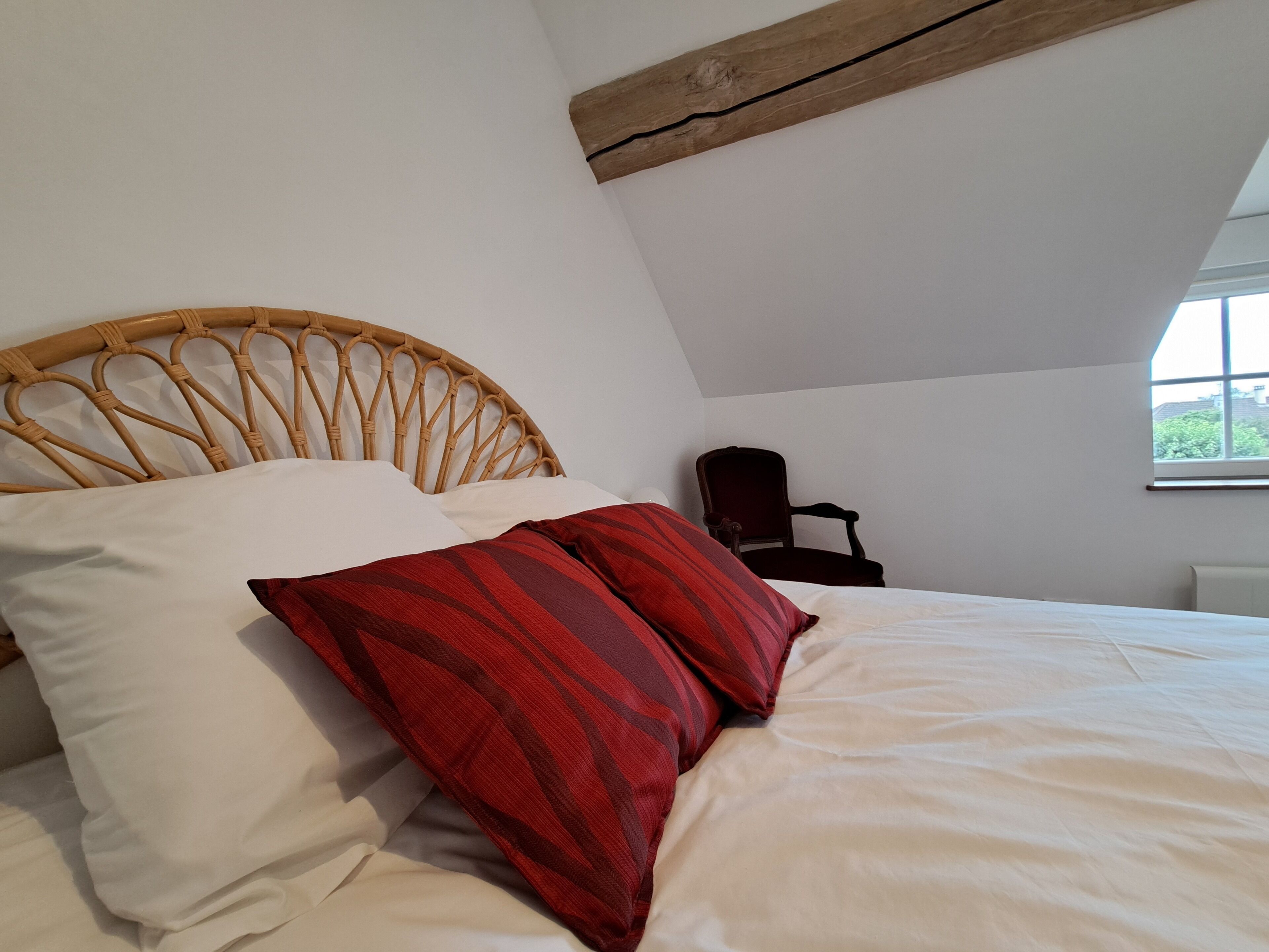 A bedroom with a wooden headboard, a bed with white and red striped pillows, a dark wooden chair, and a window with a view of greenery.