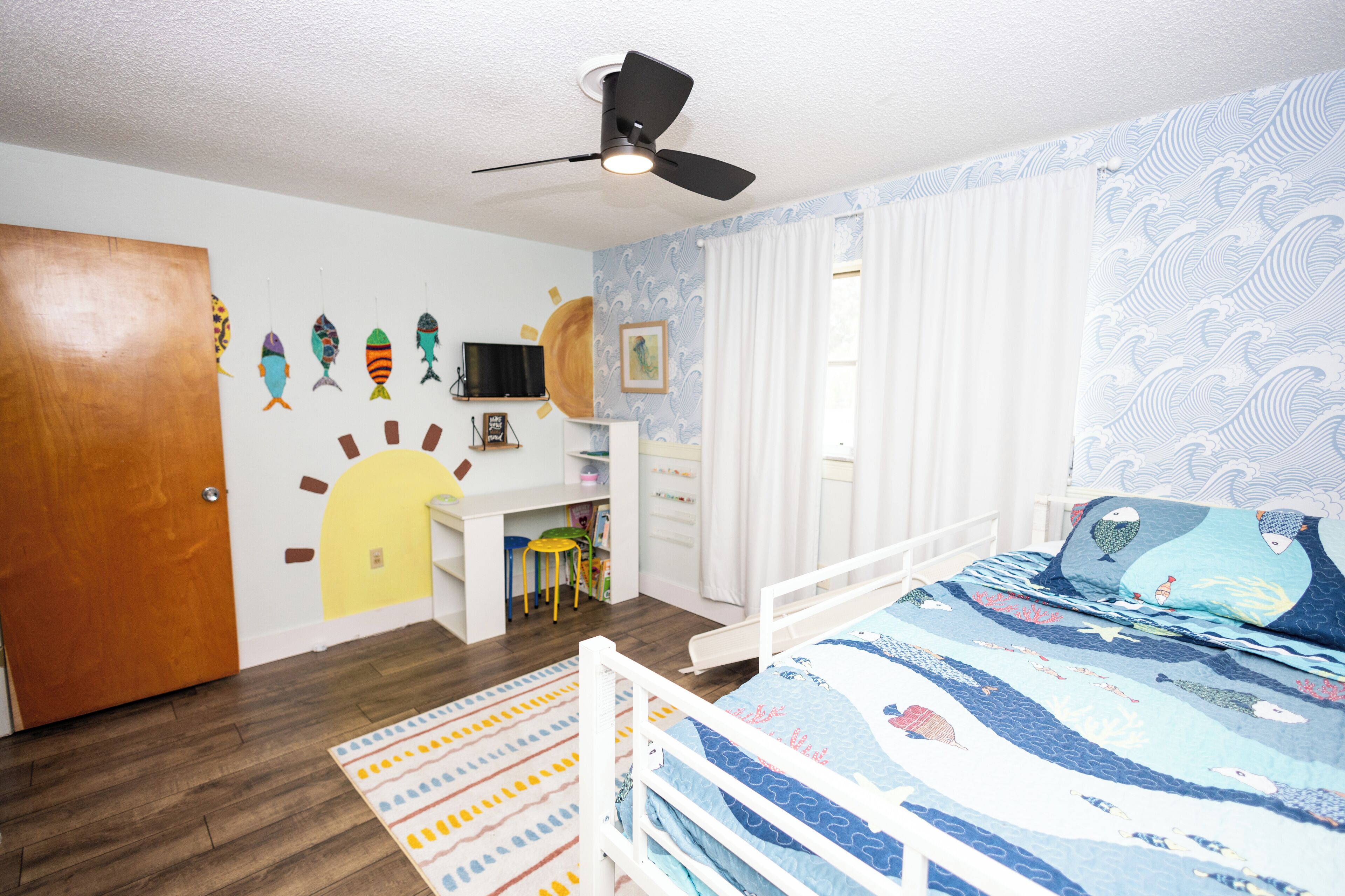 Ein Schlafzimmer mit einem Bett, einem Schreibtisch, einem Stuhl, einem Deckenventilator und einer Tür.