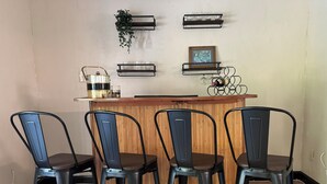 Una zona de bar con cuatro sillas de metal, un mostrador de madera y estantes con vasos y un cuadro enmarcado.