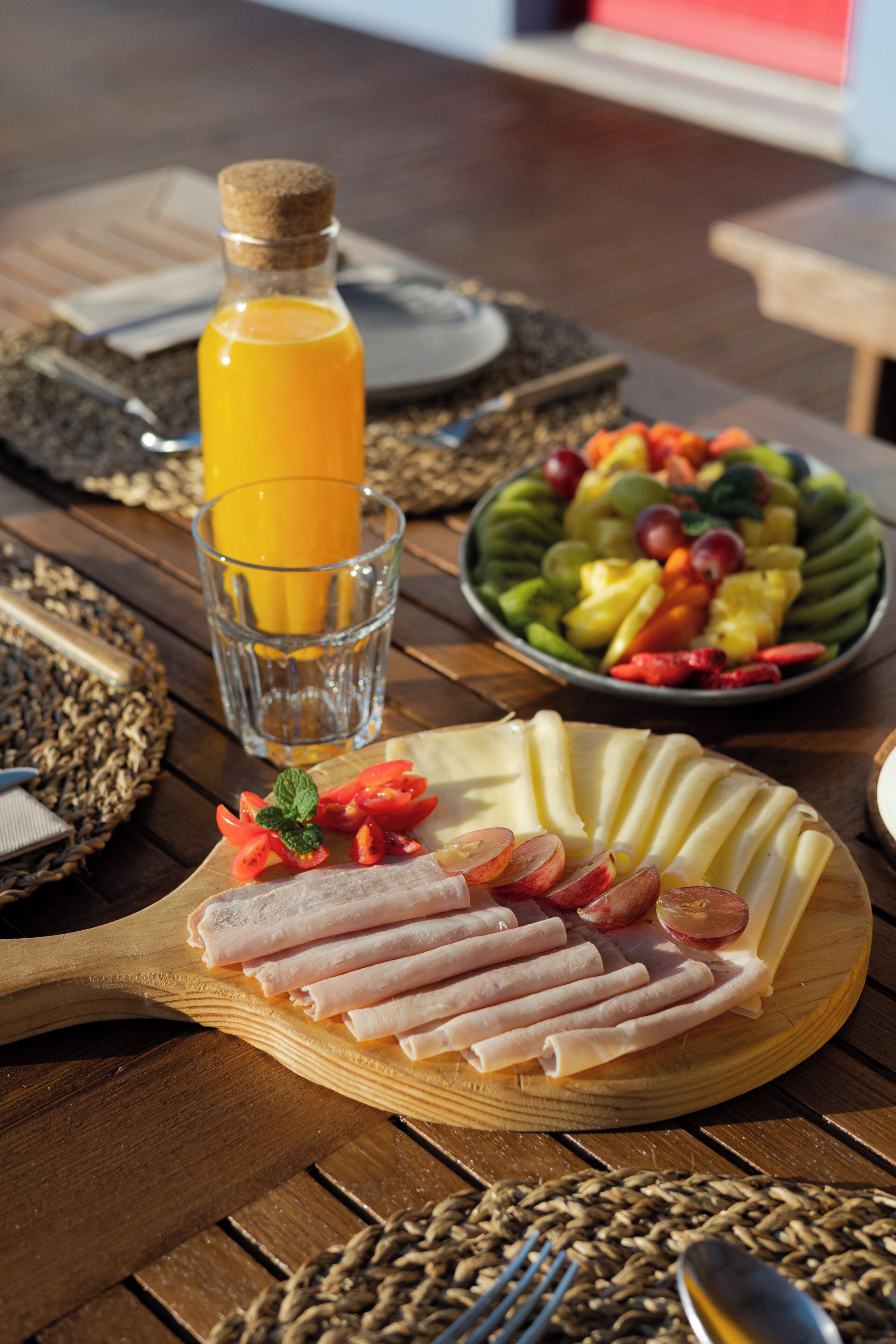 A wooden table with a plate of sliced meats, a glass of orange juice, and a bowl of fruit.
