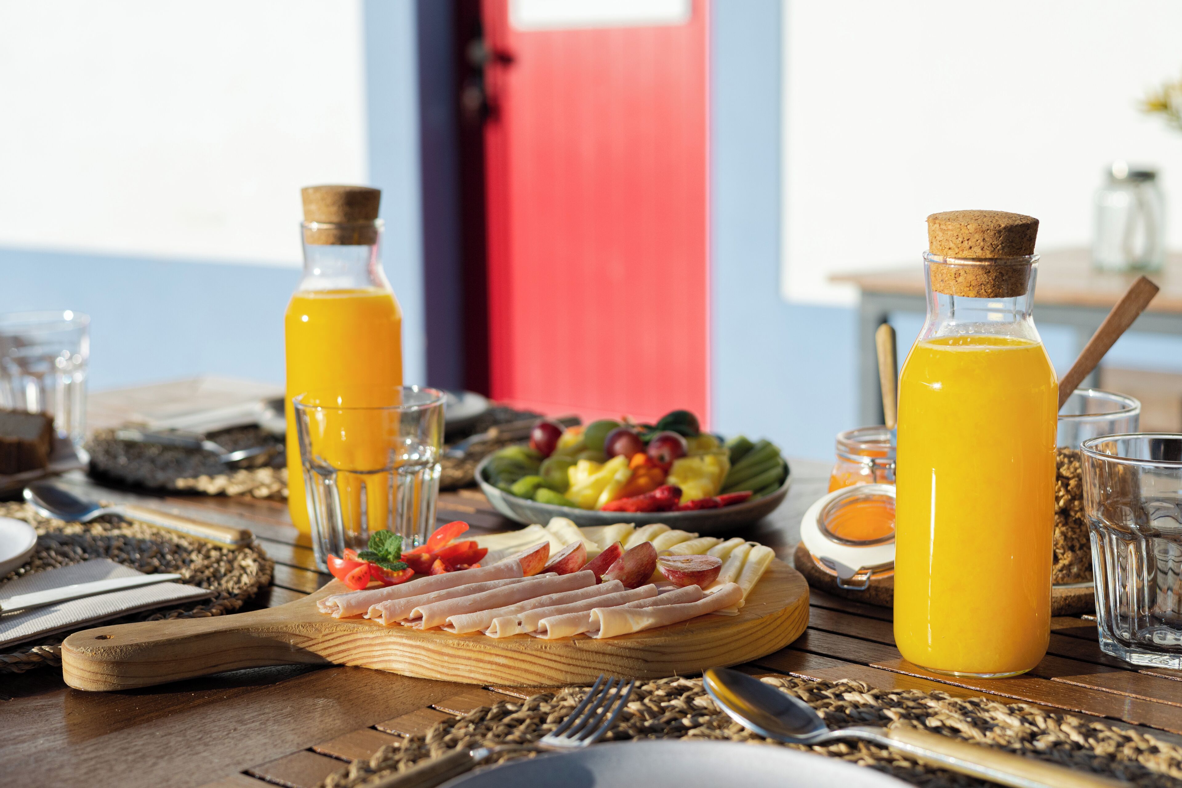 Ein Frühstückstisch mit Holzoberfläche, einer Orangensaftflasche, einer Obstschale und einer Käseplatte.