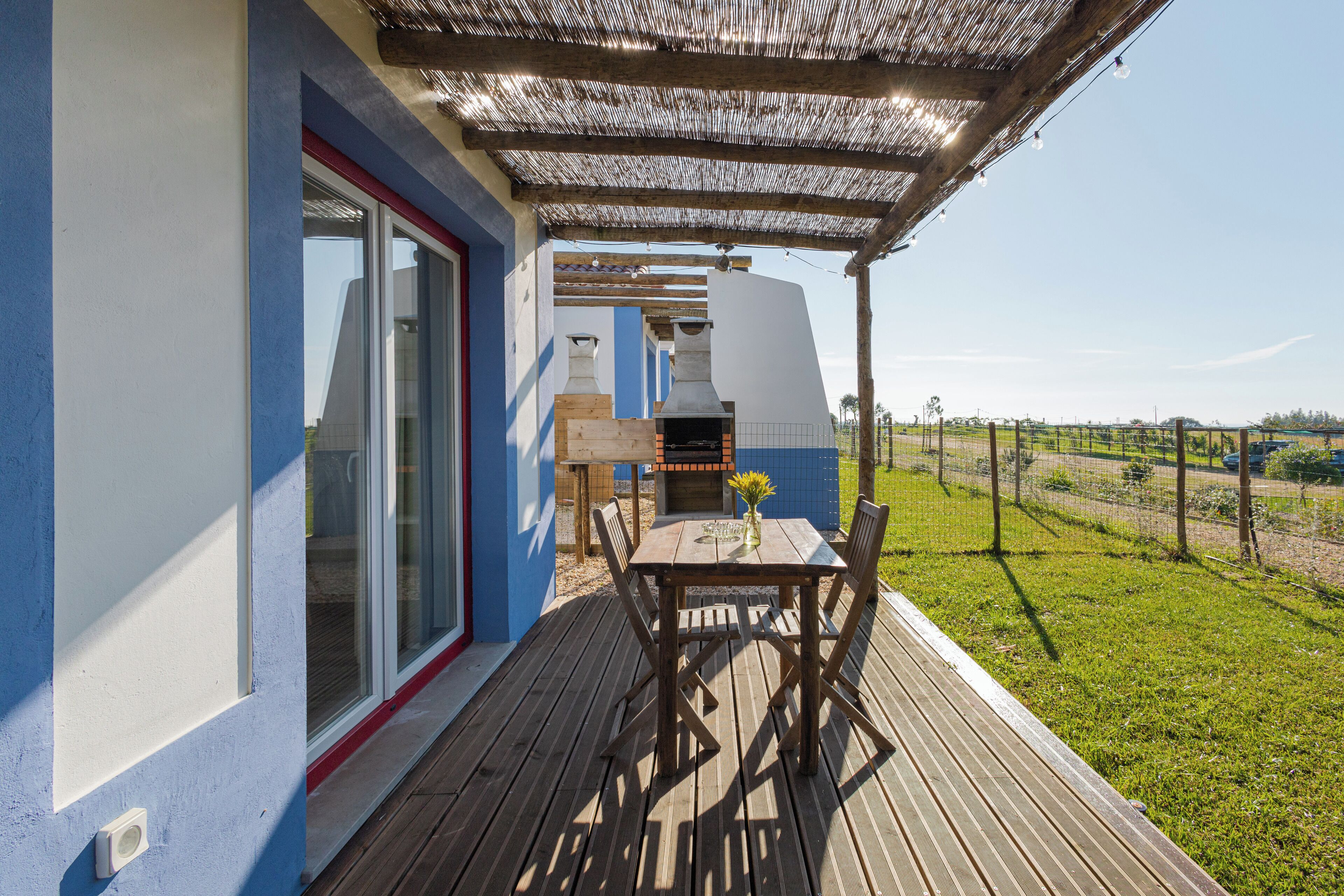 Una terraza de madera con una mesa, sillas, una parrilla y un vidrio que da a un edificio azul y blanco.