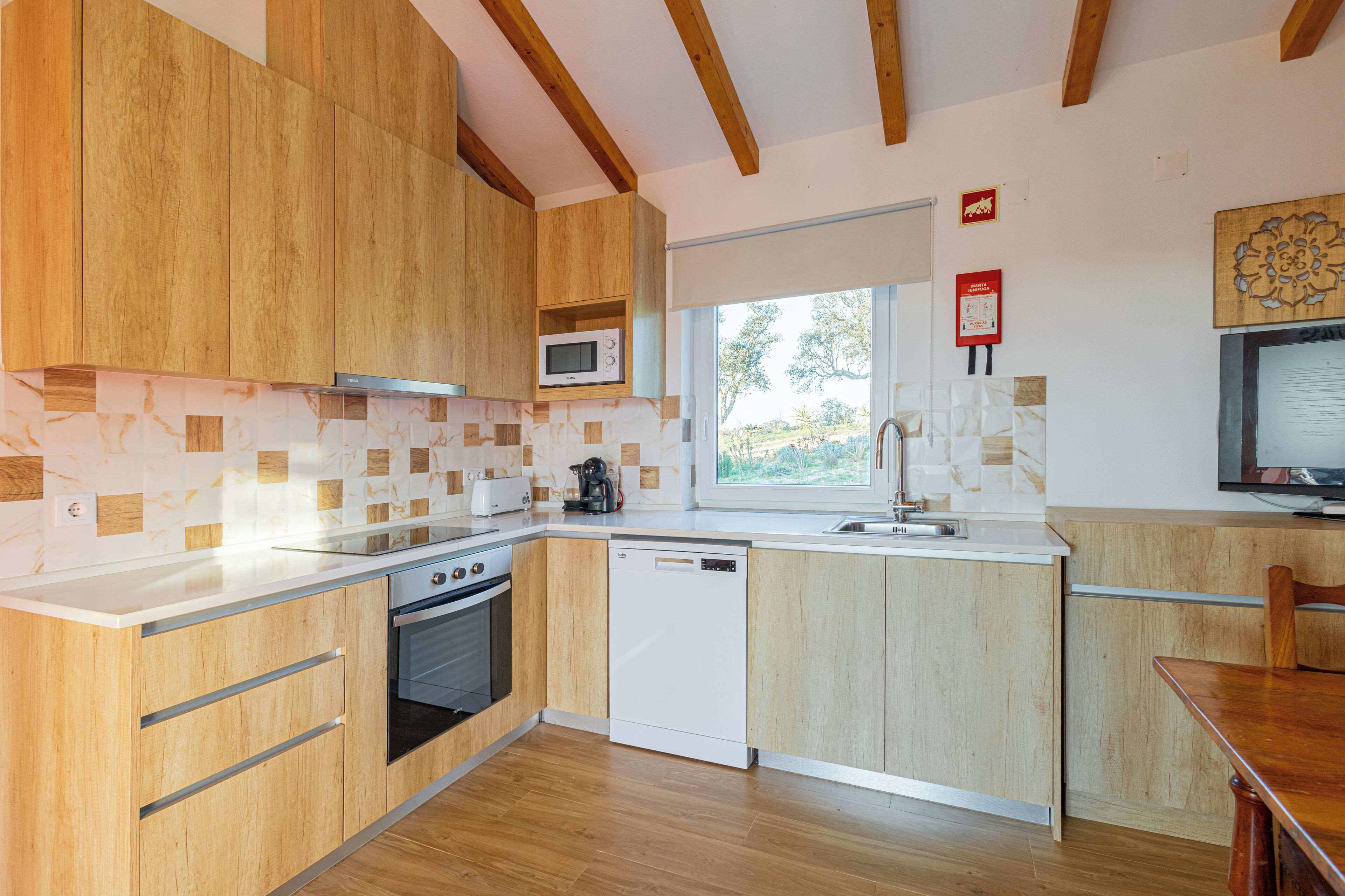 Een moderne keuken met houten kastjes, een magnetron, een vaatwasser en een raam dat voor natuurlijk licht zorgt.