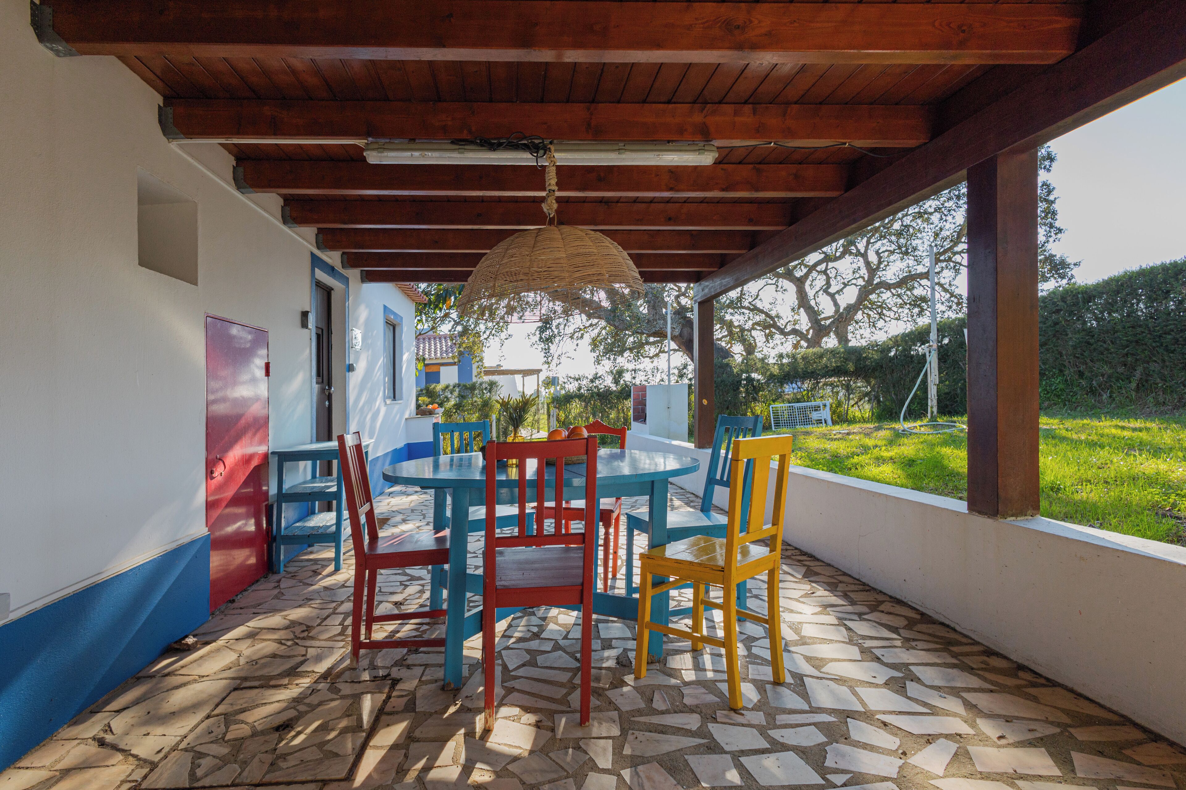 An outdoor patio with a round table and colorful chairs, a hanging light fixture, and a view of a garden.